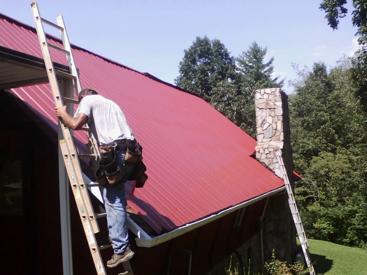 Roofer on a ladder beside a red metal roof and stone chimney — Soffit & Fascia Repair by Roofing Brothers in East New Market